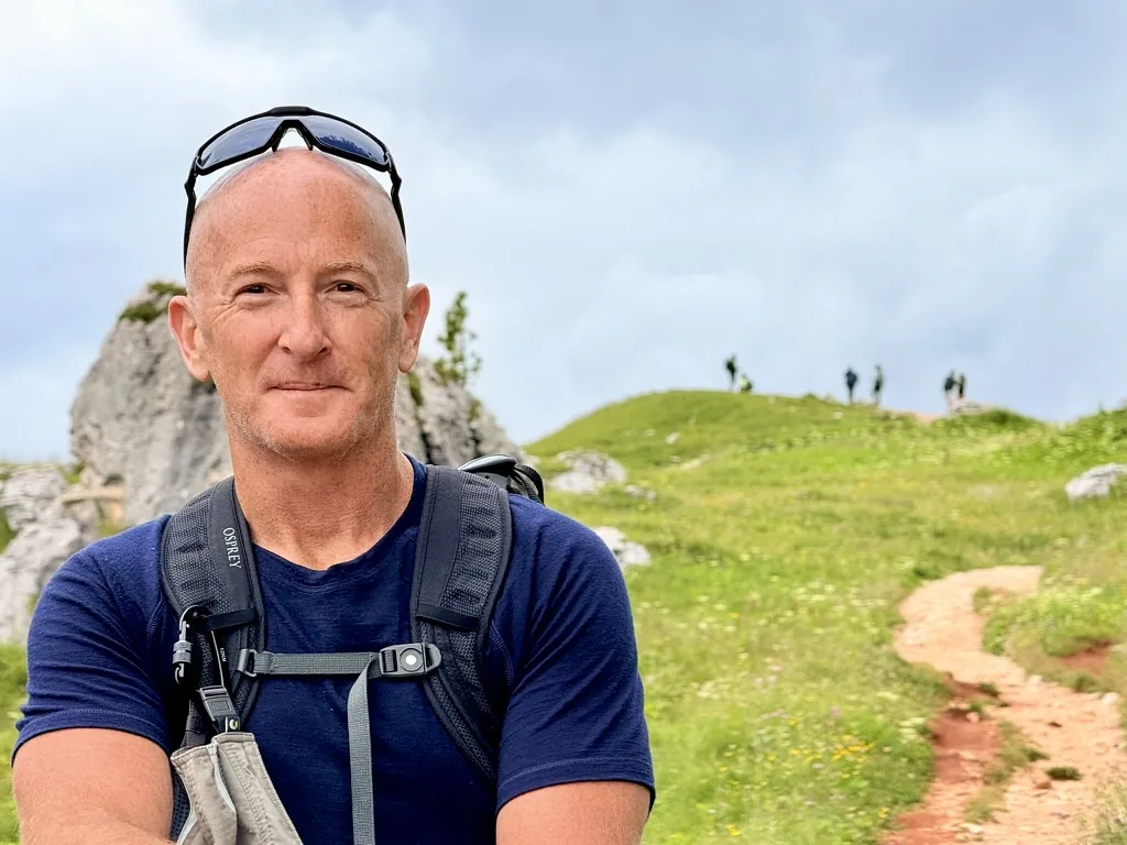 Errol Damelin hiking — navy t-shirt, Osprey backpack, green hillside.